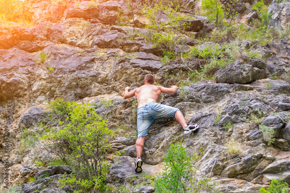 Young man climber in jeans shirts and bare torso climbs on rocks to the top in mountains of Altai without equipment and insurance