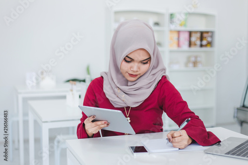 Young asian muslim woman using tablet and taking notes in notebook in a white cafe.
