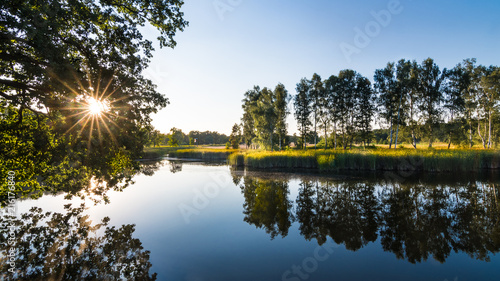 Idyllic summer landscape. Pond and sunset. South Bohemia, Europe. Blue water surface in natural scenery. Beautiful mirroring of birches and foliage of trees. Sunbeams. Green reeds on lake shore.