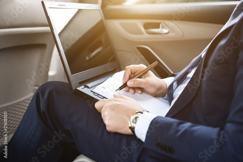 Man in a business suit write on notebook with laptop in the car