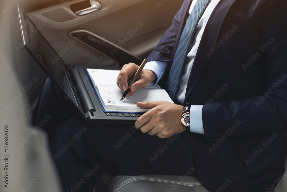 Man in a business suit write on notebook with laptop in the car Man in a business suit write on notebook with laptop in the car
