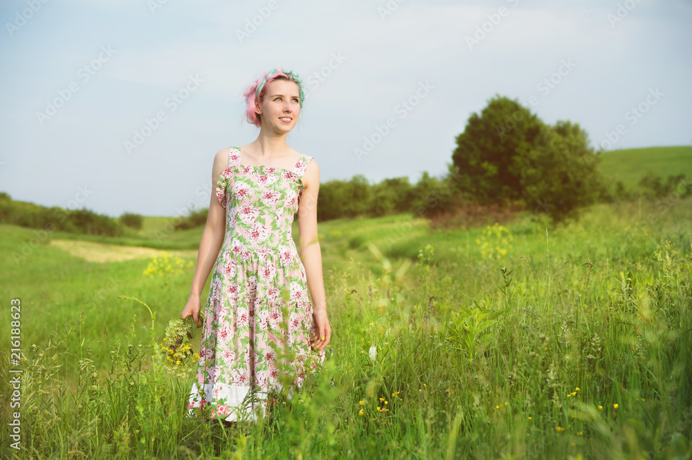 young happy smiling girl in a calico dress with a bouquet is walking along a country road with green grass young happy smiling girl in a calico dress with a bouquet is walking along a country road with green grass