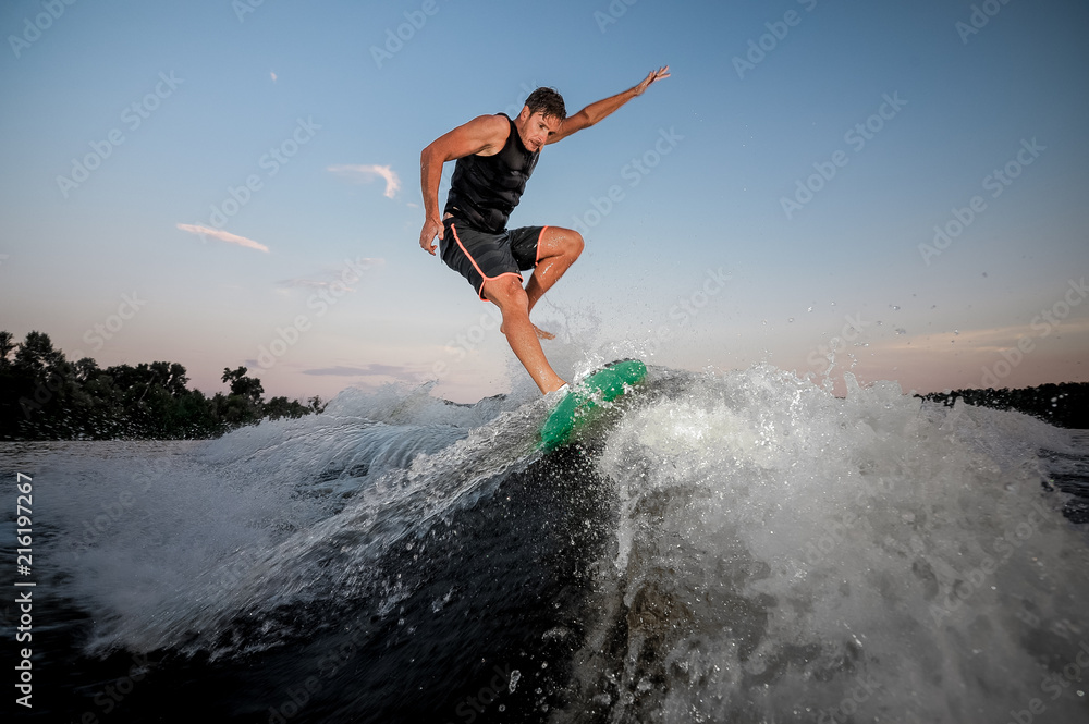Active and young man jumping on wakesurf down the river waves at the sunset