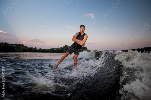 Young wakesurfer riding down the river waves crossing his hands at the sunset