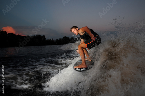 Young and active man riding on wakesurf down the river during summer sunset against the city