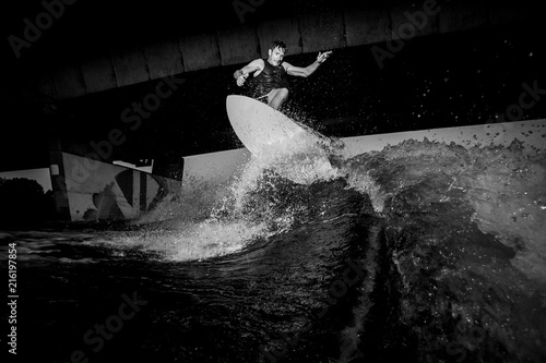 Black and white photo of active wakesurfer riding on river waves against the bridge