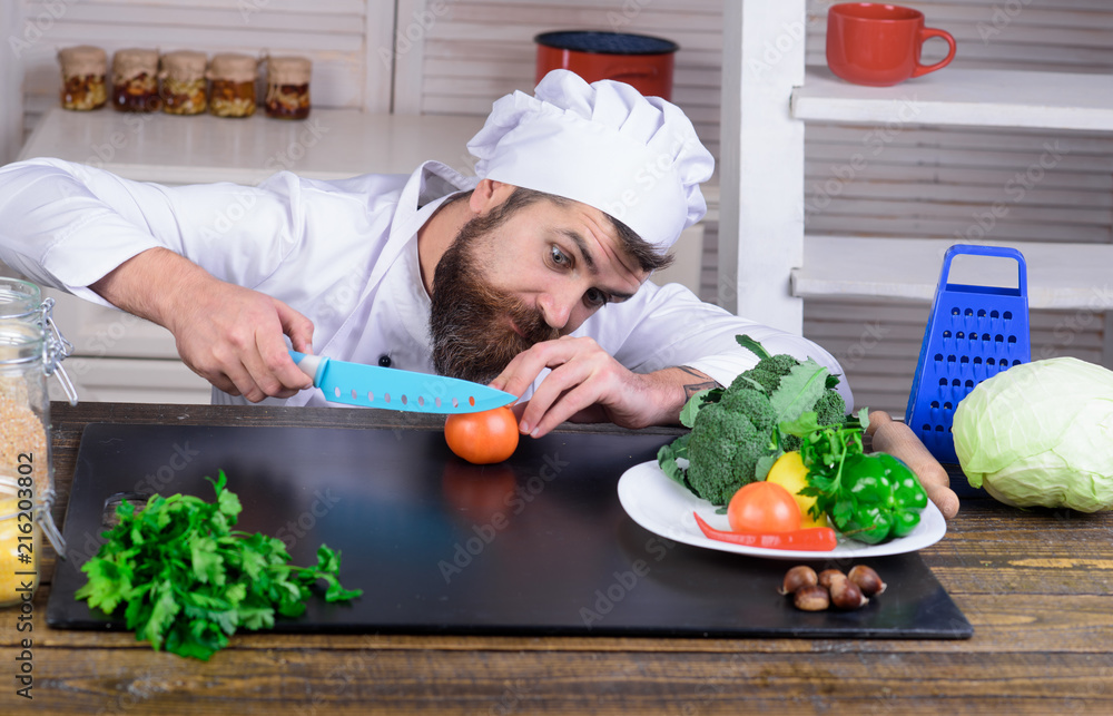 Professional male cook in white suit cut tomato with knife. Diet, healthy food, vegetarian concept - attractive bearded chef in kitchen. Preparing dishes. Organic food concept. Chef man. Master chef. Professional male cook in white suit cut tomato with knife. Diet, healthy food, vegetarian concept - attractive bearded chef in kitchen. Preparing dishes. Organic food concept. Chef man. Master chef.