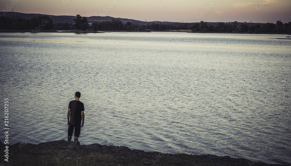 Young man standing and back to back with casual clothes on the lake shore at sunset, with eucalyptus forest in the background