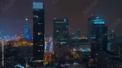 Drone footage of warsaw center at night. In the front there are modern, glazed skyscrapers, in the background the Palace of Culture and Science.