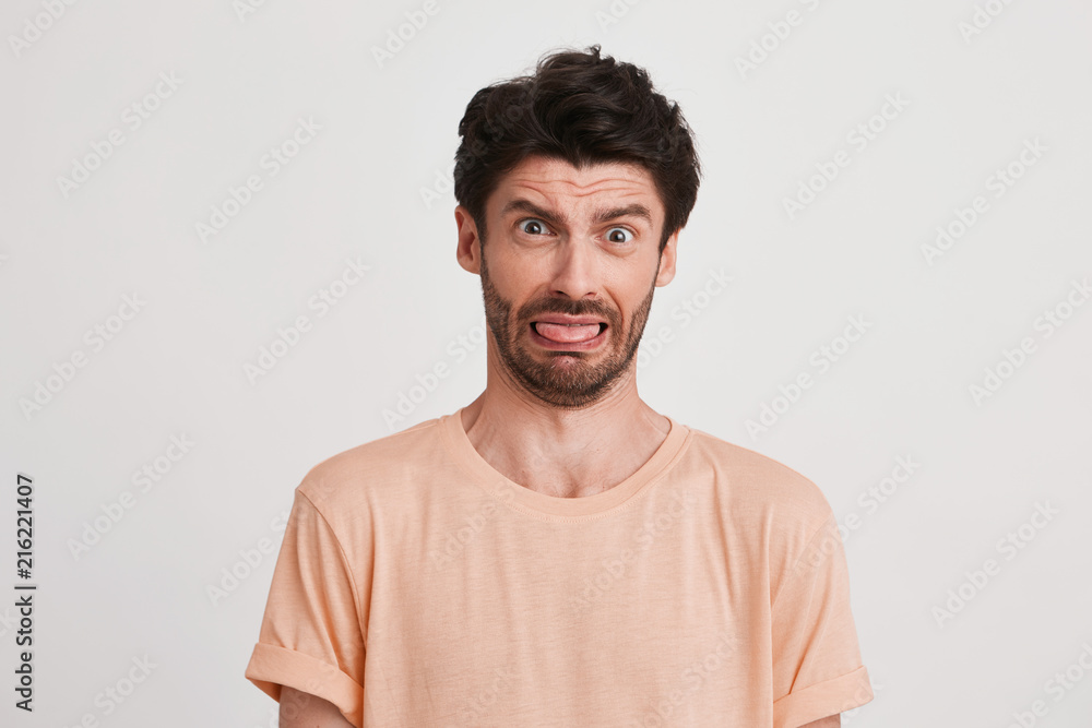 Closeup of funny dissatisfied young man with bristle wears peach t shirt looks displeased and shows tongue isolated over white background Closeup of funny dissatisfied young man with bristle wears peach t shirt looks displeased and shows tongue isolated over white background
