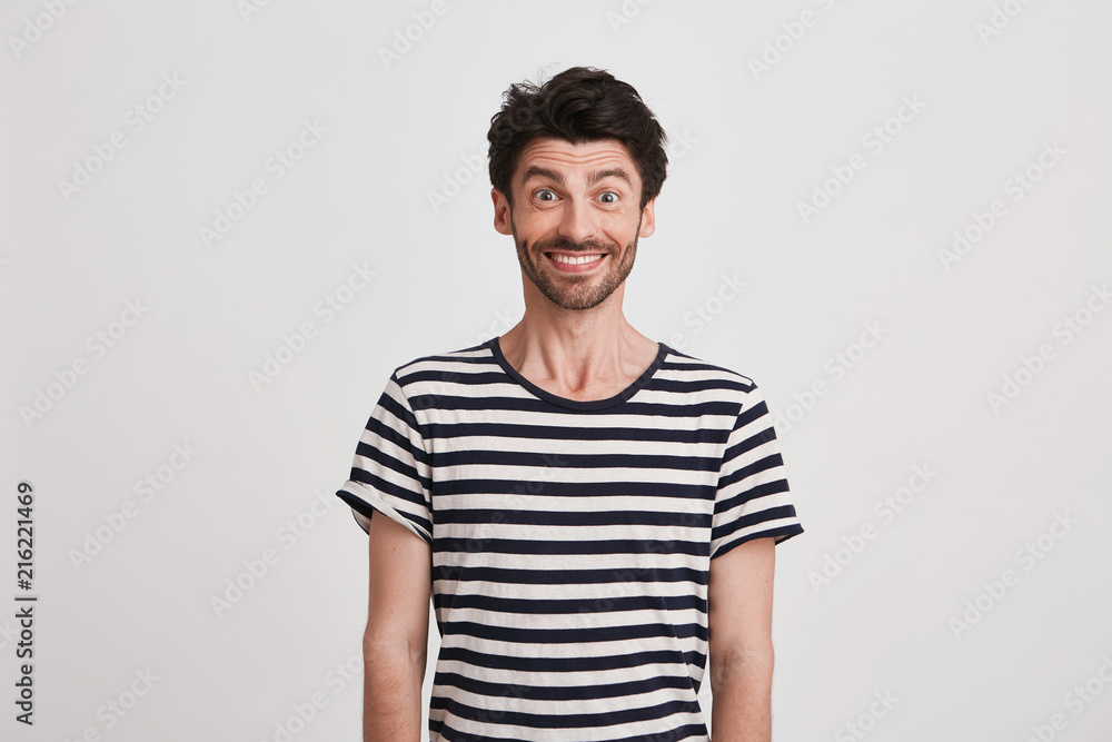 Portrait of happy attractive young man with bristle wears striped t shirt feels excited and smiling isolated over white background Looks directly in camera Portrait of happy attractive young man with bristle wears striped t shirt feels excited and smiling isolated over white background Looks directly in camera