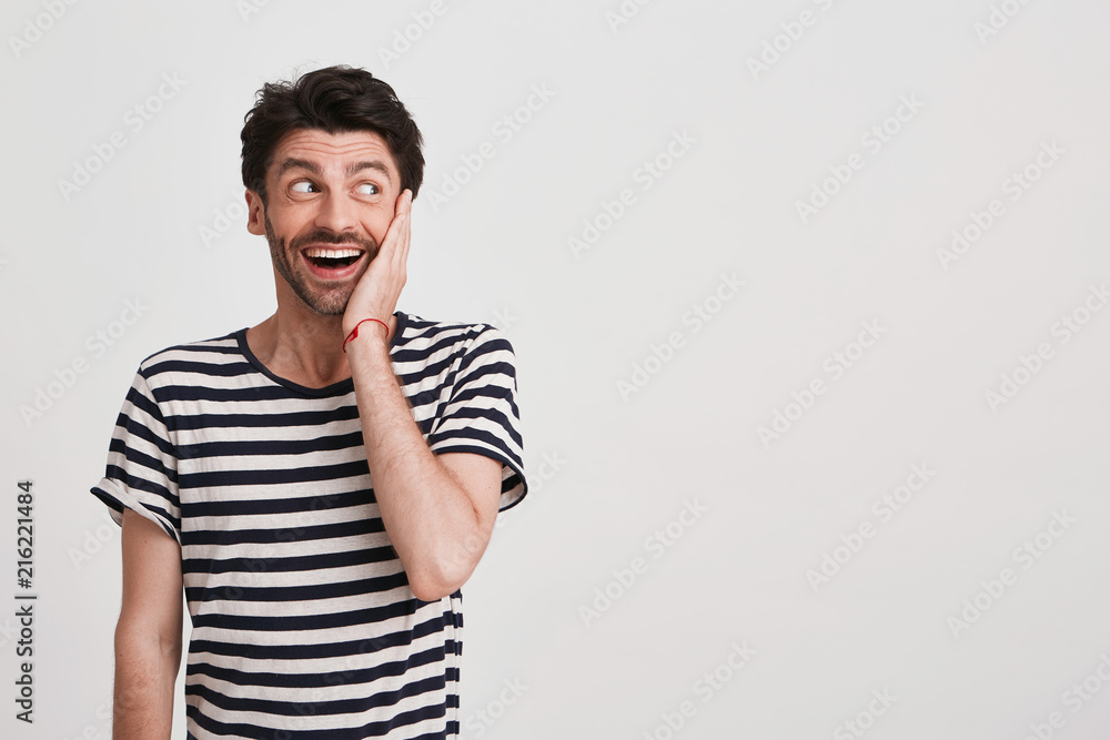 Closeup of surprised excited young man with bristle and hand on cheek wears striped t shirt feels happy and looks to the side isolated over white background Closeup of surprised excited young man with bristle and hand on cheek wears striped t shirt feels happy and looks to the side isolated over white background