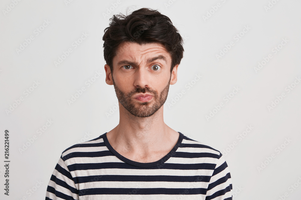 Fototapeta premium Portrait of pensive confused young man with bristle wears striped t shirt feels embarrassed and thinking isolated over white background Looks directly in camera
