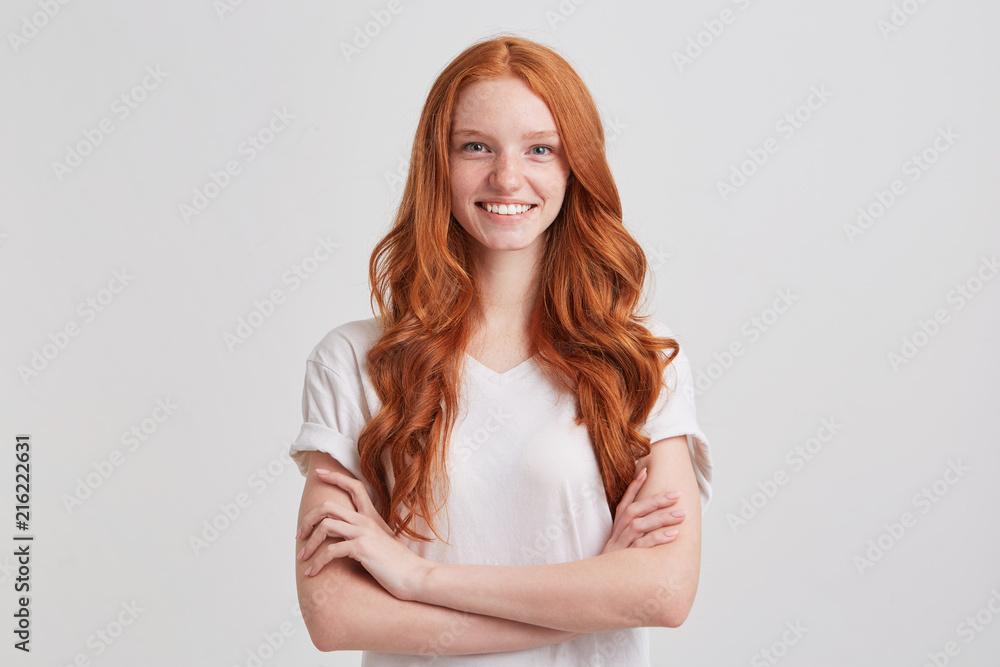 Closeup of happy confident young woman with long wavy red hair and freckles wears t shirt keeps arms crossed and looks directly in camera isolated over white background Closeup of happy confident young woman with long wavy red hair and freckles wears t shirt keeps arms crossed and looks directly in camera isolated over white background