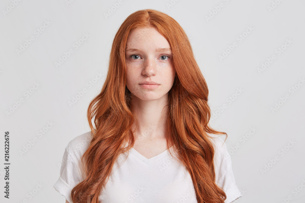 Closeup of serious beautiful redhead young woman with long wavy hair and freckles wears t shirt feels confident and looks directly in camera isolated over white background Closeup of serious beautiful redhead young woman with long wavy hair and freckles wears t shirt feels confident and looks directly in camera isolated over white background
