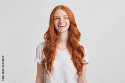 Portrait of cheerful cute redhead young woman with long wavy hair and freckles wears stylish t shirt looks relaxed and laughing isolated over white background Feels happy photo