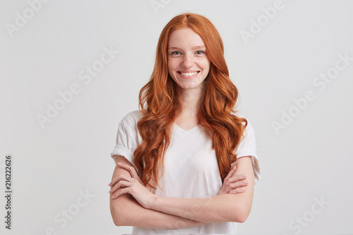 Closeup of smiling lovely redhead young woman with long wavy hair and freckles wears stylish t shirt looks confident and keeps arms crossed isolated over white background photo