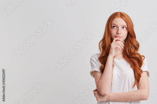 Closeup of cute thoughtful redhead young woman with long wavy hair and freckles wears stylish t shirt thibnking and planning her working day isolated over white background
