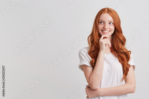 Portrait of happy charming redhead young woman with long wavy hair and freckles wears t shirt looks to the side, thinks and plans her birthday party isolated over white background