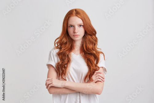 Portrait of serious beautiful redhead young woman with long wavy hair and freckles wears t shirt keeps arms crossed and feels confident isolated over white background