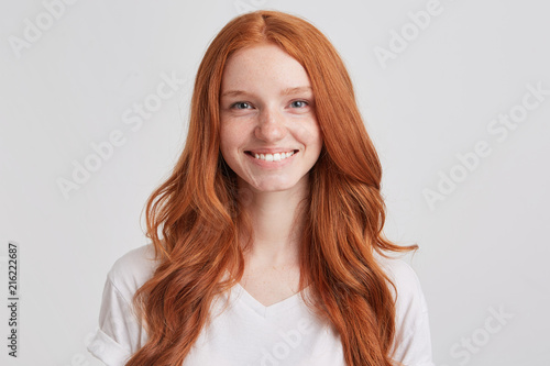 Closeup of cheerful lovely redhead young woman with long wavy hair and freckles wears t shirt looks confident and feels happy isolated over white background Looks directly in camera photo