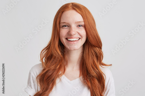 Portrait of cheerful pretty redhead young woman with long wavy hair and freckles wears t shirt feels joyful and laughing isolated over white background Looks directly in camera