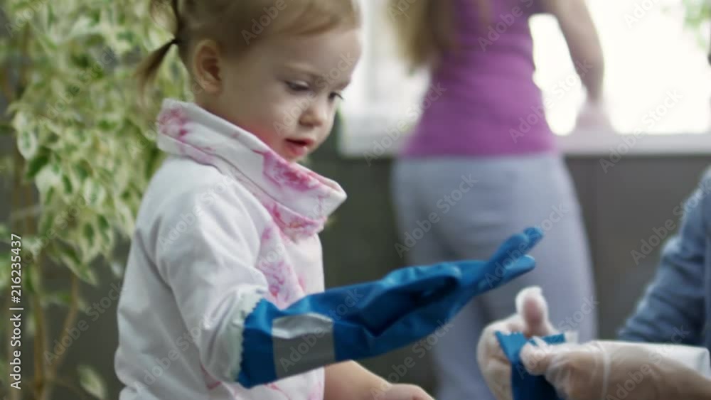 Handheld medium shot of happy young mother putting on cleaning gloves on cute little girl and giving her microfiber cloth Handheld medium shot of happy young mother putting on cleaning gloves on cute little girl and giving her microfiber cloth