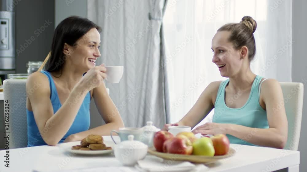 PAN of happy young female couple chatting and drinking tea in kitchen PAN of happy young female couple chatting and drinking tea in kitchen