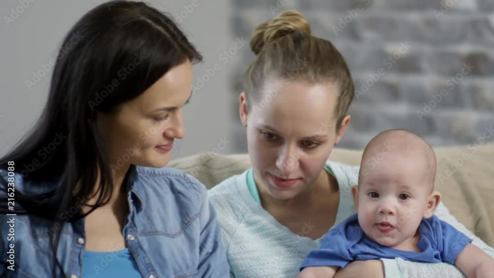 Handheld medium shot of happy female couple chatting and interacting with cute baby boy while reading book on sofa Handheld medium shot of happy female couple chatting and interacting with cute baby boy while reading book on sofa