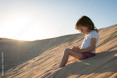 Little girl sitting on sand dune and playing game on phone.