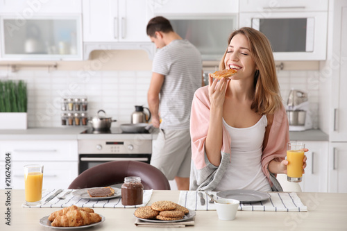 Beautiful young woman having breakfast while her beloved boyfriend cooking in kitchen