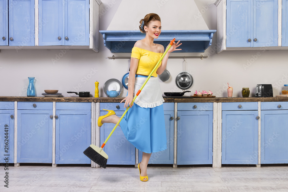 Retro / pin up girl female woman / housewife wearing colorful top, skirt and white apron holding mop and cleaning washing floor in the kitchen with blue cabinets and utensils. Housework concept Retro / pin up girl female woman / housewife wearing colorful top, skirt and white apron holding mop and cleaning washing floor in the kitchen with blue cabinets and utensils. Housework concept
