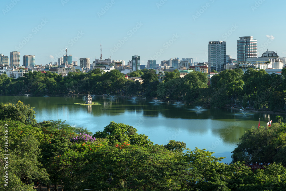 Fototapeta premium Hoan Kiem lake or Sword lake, Ho Guom in Hanoi, Vietnam with Turtle Tower, green trees and buildings on horizon