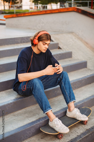 Young skater in orange headphones thoughtfully using cellphone sitting on stairs with skateboard at skate park © Anton