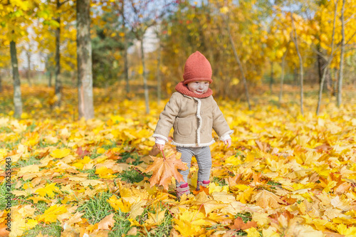 Little smiling girl in warm cap with maple leaf play in autumn forest