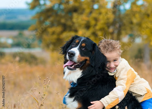 cute boy in yellow jacket hugging his dog on walk through autumn meadow Berner Sennenhund