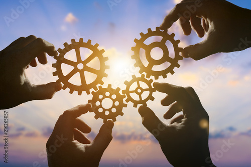 Four hands of businessmen collect gear from the gears of the details of puzzles. against the backdrop of dramatic sunlight. The concept of a business idea. Teamwork, strategy, cooperation, creativity photo