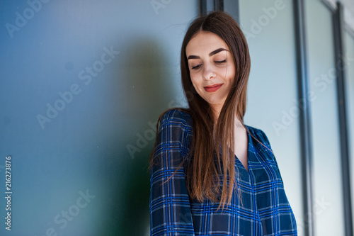 Portrait of young brunette girl in blue checkered shirt against large windows.