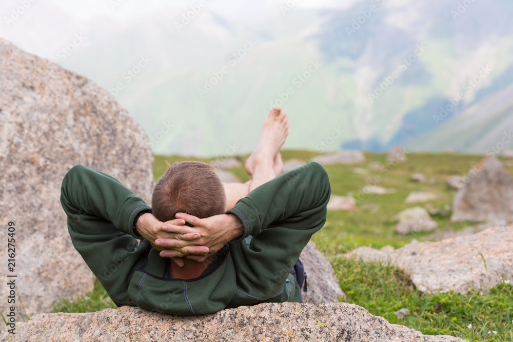 a man with hands behind his head is lying on a stone in highland relaxing and looking into the distance at the mountains