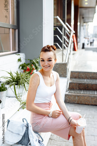 Positive summer concept. Cute cheerful girl in a white t-shirt and pink skirt drinks a milkshake or soft drink on the background of a summer cafe. She smiles friendly at the camera