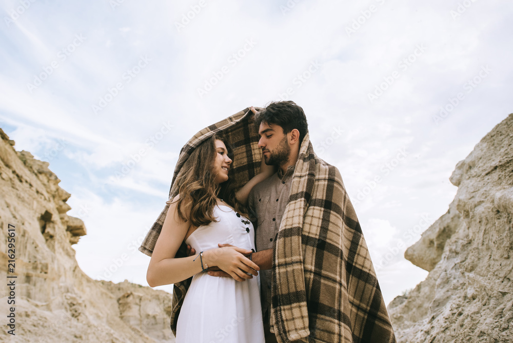 Fototapeta premium happy couple embracing under blanket in sand canyon with cloudy sky