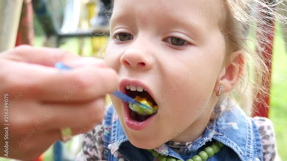 Face portrait mom feeds her cute child girl from a spoon with boiled grains of corn