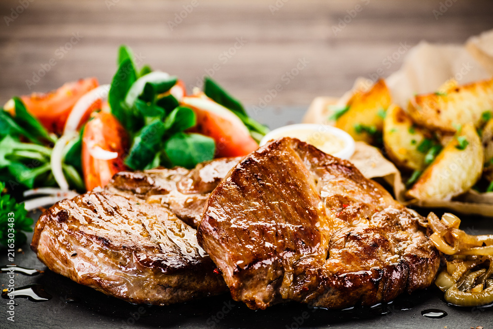 Grilled steaks with baked potatoes and vegetables served on black stone on wooden table 