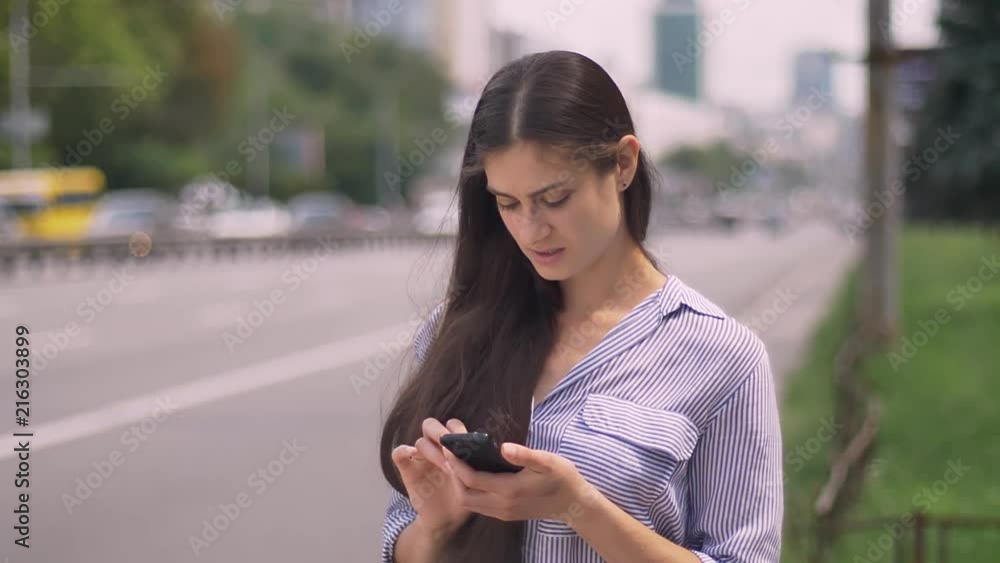 Beautiful young businesswoman typing on phone and waiting for taxi, standing on street near road Beautiful young businesswoman typing on phone and waiting for taxi, standing on street near road