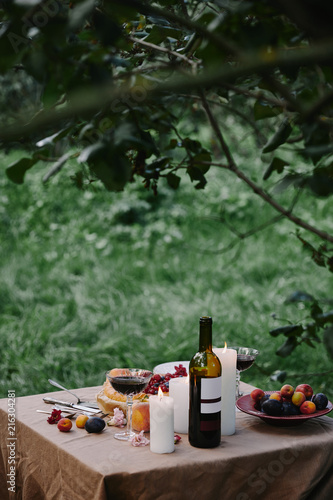candles, wine bottle and fruits on table in garden for dinner