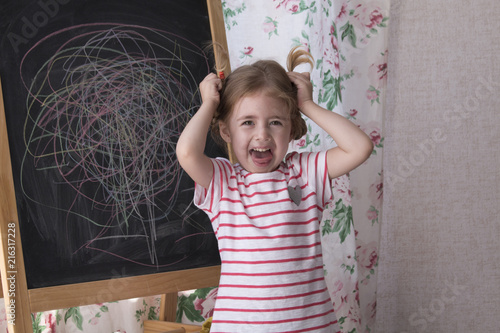 Child is drawing with pieces of color chalk on the chalk board. Girl is expressing creativity and looking at the camera, smiling playroom. Concept of expression and learning