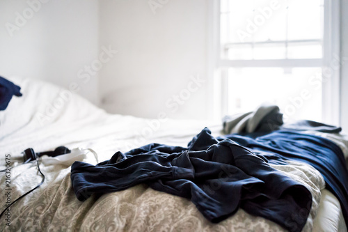 Messy room with dark blue black clothes lying on top of bed sheets comforter in depressing cold cool room closeup with window, headphones, bedroom