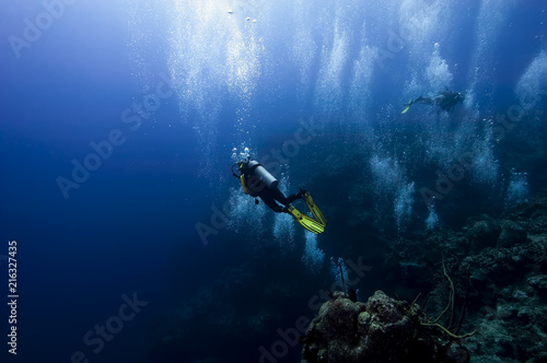 A group of divers float at the top of the 1000ft drop wall in Grand cayman whilst they do a safety rest stop after a deep dive. Multitudes of bubbles are cascading upwards to the surface of the ocean.
