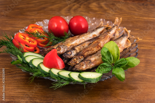 Fried capelin on a round glass plate with cucumber slices, sweet pepper circles, dill sprigs, green basil leaves and red whole tomatoes.