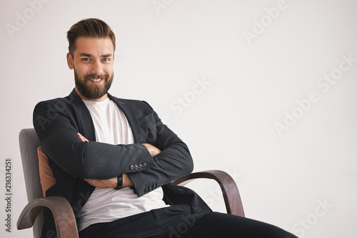 Business, career and success concept. Cheerful stylish young unshaven businessman with happy smile, sitting in armchair against blank studio wall background with copy space for your information photo